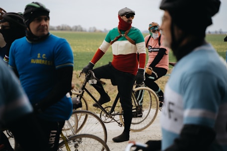Cyclists are gathered on a road with vintage-style bicycles, wearing colorful outfits that suggest a retro theme. The participants appear to be in a relaxed setting with an overcast sky, indicating a cycling event in a rural area with green fields in the background.