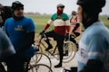 Cyclists are gathered on a road with vintage-style bicycles, wearing colorful outfits that suggest a retro theme. The participants appear to be in a relaxed setting with an overcast sky, indicating a cycling event in a rural area with green fields in the background.