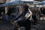 A person wearing vintage cycling attire with an armband numbered 41 is riding a bicycle. The background features a booth with Bianchi branding and various bicycle frames and wheels on display. The setting seems to be an outdoor event or market with people milling about.