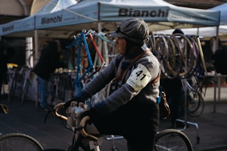 A person wearing vintage cycling attire with an armband numbered 41 is riding a bicycle. The background features a booth with Bianchi branding and various bicycle frames and wheels on display. The setting seems to be an outdoor event or market with people milling about.