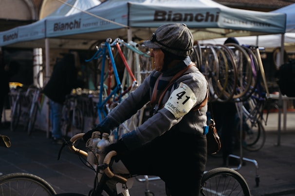 A person wearing vintage cycling attire with an armband numbered 41 is riding a bicycle. The background features a booth with Bianchi branding and various bicycle frames and wheels on display. The setting seems to be an outdoor event or market with people milling about.
