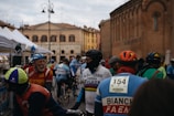 A group of cyclists gathered in an outdoor area of an old town square, surrounded by historic buildings. They are wearing colorful cycling gear with vintage designs, including helmets and jerseys. A variety of tents are visible in the background, suggesting an event or race is taking place.