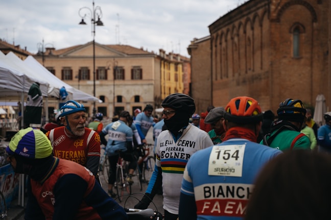 A group of cyclists gathered in an outdoor area of an old town square, surrounded by historic buildings. They are wearing colorful cycling gear with vintage designs, including helmets and jerseys. A variety of tents are visible in the background, suggesting an event or race is taking place.