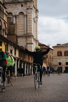 Cyclists ride along a cobblestone street beside historic architecture with arches and columns. A person in a green helmet is at the forefront, raising both hands in a peace sign gesture. The background features a tall building with intricate stonework.