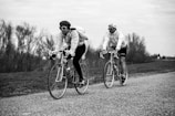 Two cyclists are riding vintage-style road bikes along a countryside path. Both individuals wear sunglasses and cycling caps, with one cyclist carrying a backpack. The background features leafless trees under an overcast sky, creating a serene and nostalgic atmosphere.