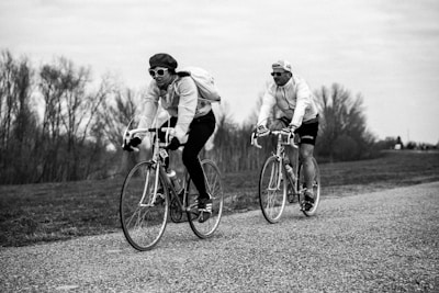 Two cyclists are riding vintage-style road bikes along a countryside path. Both individuals wear sunglasses and cycling caps, with one cyclist carrying a backpack. The background features leafless trees under an overcast sky, creating a serene and nostalgic atmosphere.