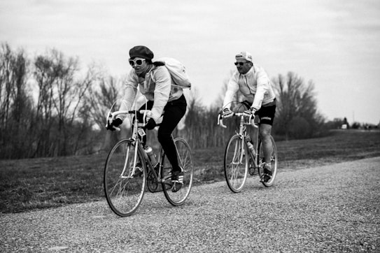 Two cyclists are riding vintage-style road bikes along a countryside path. Both individuals wear sunglasses and cycling caps, with one cyclist carrying a backpack. The background features leafless trees under an overcast sky, creating a serene and nostalgic atmosphere.