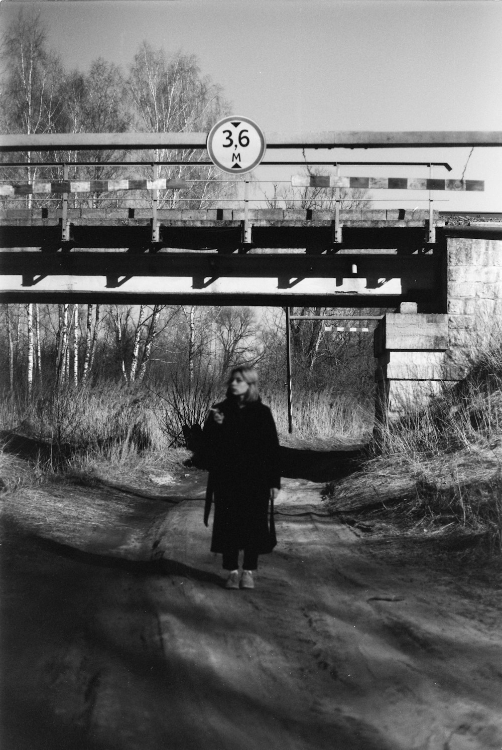 a woman walking down a dirt road under a bridge