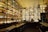 A bar with shelves filled with colorful bottles, illuminated from behind. The ceiling features ceiling fans, and there are two people at the bar counter. The ambiance is warm with a vintage feel, highlighting the bottles on display.