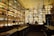 A bar with shelves filled with colorful bottles, illuminated from behind. The ceiling features ceiling fans, and there are two people at the bar counter. The ambiance is warm with a vintage feel, highlighting the bottles on display.