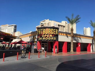 A restaurant with a large sign reading 'Fatburger Open 24 Hours' against a bright blue sky. The building has red accents and is surrounded by palm trees. In the background, tall hotel buildings are visible. Red umbrellas cover tables outside, and a few people are near the entrance.