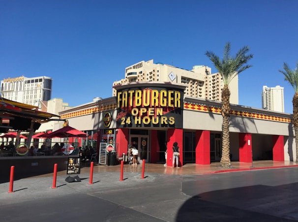 A restaurant with a large sign reading 'Fatburger Open 24 Hours' against a bright blue sky. The building has red accents and is surrounded by palm trees. In the background, tall hotel buildings are visible. Red umbrellas cover tables outside, and a few people are near the entrance.