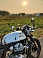 A Royal Enfield motorcycle is parked on a rural road with an expansive, grassy field in the background. The sun is setting or rising, adding a warm glow to the scene. Palm trees and distant buildings are visible along the horizon.