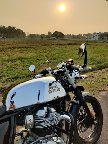 A Royal Enfield motorcycle is parked on a rural road with an expansive, grassy field in the background. The sun is setting or rising, adding a warm glow to the scene. Palm trees and distant buildings are visible along the horizon.