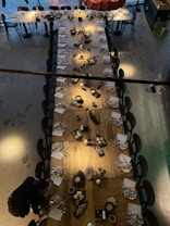 A long dining table is set with elegant tableware, including plates, glassware, and neatly arranged napkins. Assorted platters of appetizers and canap&eacute;s are placed along the center, flanked by candles providing ambient lighting. Surrounding the table are black chairs, and a person is seen taking a photograph at one end of the table. The floor has a polished, industrial look, and there's a modern aesthetic to the overall scene.