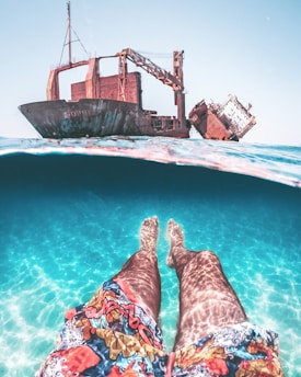 A split-level view combining underwater and above-water perspectives. Visible underwater are a person's legs in colorful swim shorts and clear blue water. Above the waterline, a large, rusty shipwreck is prominently featured, partially tilted, against a clear sky.