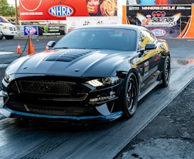 A sleek black sports car is positioned on a drag racing track, minutes away from a potential race. The car has a shiny, polished exterior with aerodynamic features and a prominent front grille. In the background, there are advertisements for various racing and automotive brands, along with an NHRA banner. Orange traffic cones are visible near the track.