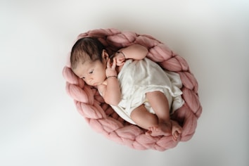 A newborn baby is resting comfortably inside a soft, knitted pink basket. The baby is wrapped in a white cloth and is looking to the side, appearing relaxed and peaceful.