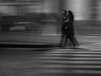A blurred image capturing a couple walking across a zebra crossing at night. The motion blur gives the scene a sense of movement and speed while the image remains monochromatic, adding a timeless and atmospheric quality.