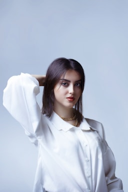 A young woman with dark hair is wearing a white blouse and gold necklace. Her hair is styled, and she has a confident expression on her face. The background is a soft, light blue.
