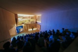 A group of people is sitting in a darkened room with wooden walls, watching a large screen where a presentation or film is being projected. The audience is seated on tiered steps, and the environment has a modern, minimalist design, with dim blue lighting creating a calm atmosphere and a potted plant near the screen adding a touch of nature.