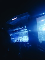 A large outdoor concert stage illuminated with blue lights, featuring a musician or performer in front of a cheering crowd. The stage has an expansive display screen showing graphics and the audience is visible in silhouette.