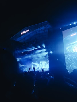 A large outdoor concert stage illuminated with blue lights, featuring a musician or performer in front of a cheering crowd. The stage has an expansive display screen showing graphics and the audience is visible in silhouette.