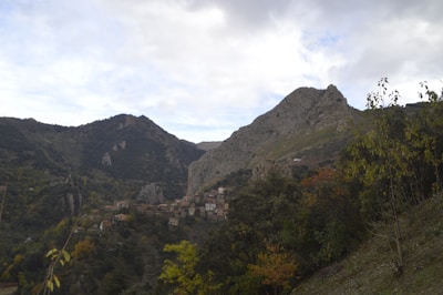 A picturesque village nestled in a mountainous landscape, surrounded by lush greenery and trees. The village consists of small, rustic houses with earthy tones, scattered along the hillside. Majestic mountains rise in the background under a partially cloudy sky, creating a serene and natural setting.