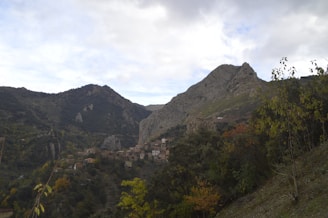 A picturesque village nestled in a mountainous landscape, surrounded by lush greenery and trees. The village consists of small, rustic houses with earthy tones, scattered along the hillside. Majestic mountains rise in the background under a partially cloudy sky, creating a serene and natural setting.