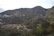 A picturesque hillside landscape featuring numerous houses scattered among dense green trees. The buildings are nestled within lush forested hills, and a smoky haze is visible in the distance. The mountains in the background are rugged and covered with a mix of vegetation.