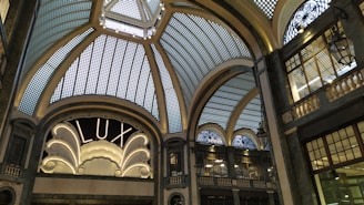 A grand, ornate interior with an elaborate glass ceiling composed of geometric panels, adorned with decorative metalwork. An elegant sign reading 'LUX' is illuminated, framed by intricate architectural details. Dim lighting casts soft shadows, enhancing the sophisticated ambiance.