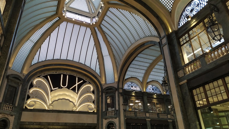 A grand, ornate interior with an elaborate glass ceiling composed of geometric panels, adorned with decorative metalwork. An elegant sign reading 'LUX' is illuminated, framed by intricate architectural details. Dim lighting casts soft shadows, enhancing the sophisticated ambiance.