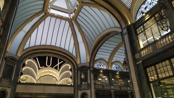 A grand, ornate interior with an elaborate glass ceiling composed of geometric panels, adorned with decorative metalwork. An elegant sign reading 'LUX' is illuminated, framed by intricate architectural details. Dim lighting casts soft shadows, enhancing the sophisticated ambiance.