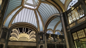 A grand, ornate interior with an elaborate glass ceiling composed of geometric panels, adorned with decorative metalwork. An elegant sign reading 'LUX' is illuminated, framed by intricate architectural details. Dim lighting casts soft shadows, enhancing the sophisticated ambiance.