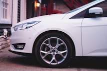 A white car is parked close to a building with a brick exterior. The focus is on the front wheel and part of the front body of the car, highlighting its sleek design. A warm light from a lamp is visible in the background, and red stairs are partially seen, adding depth to the scene.
