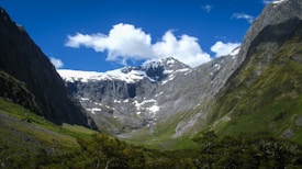 A majestic mountain range with snow-capped peaks against a vibrant blue sky. The landscape is framed by steep, rocky cliffs on both sides and lush greenery at the base.