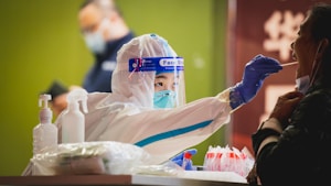 A healthcare worker wearing a protective face shield, mask, and full-body suit is administering a COVID-19 test by taking a nasal or throat swab from a patient. The setting includes various medical supplies like bottles and gloves on a table.