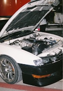 A white car with an open hood revealing its engine components is parked on a red and gray surface. The car has shiny metallic wheels and a black front bumper. In the background, part of another vehicle and a person partially visible are present.