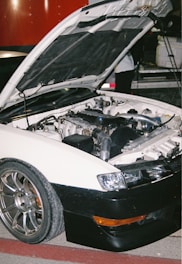 A white car with an open hood revealing its engine components is parked on a red and gray surface. The car has shiny metallic wheels and a black front bumper. In the background, part of another vehicle and a person partially visible are present.