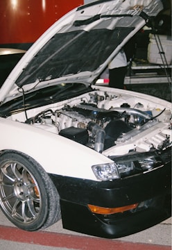 A white car with an open hood revealing its engine components is parked on a red and gray surface. The car has shiny metallic wheels and a black front bumper. In the background, part of another vehicle and a person partially visible are present.