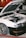A white car with an open hood revealing its engine components is parked on a red and gray surface. The car has shiny metallic wheels and a black front bumper. In the background, part of another vehicle and a person partially visible are present.