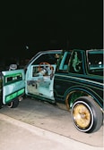 A customized classic car with its door open, revealing an interior featuring teal upholstery and a wooden steering wheel. The car's exterior is painted a dark green with intricate decorative designs and pinstriping in lighter colors. The lowrider is equipped with gold-accented rims and whitewall tires, parked on a street at night.