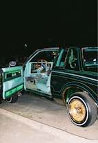 A customized classic car with its door open, revealing an interior featuring teal upholstery and a wooden steering wheel. The car's exterior is painted a dark green with intricate decorative designs and pinstriping in lighter colors. The lowrider is equipped with gold-accented rims and whitewall tires, parked on a street at night.