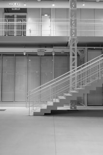 A modern, minimalistic interior space featuring a prominent staircase with sleek metal railings. The setting includes a high ceiling, large glass panels, and visible structural beams, conveying a sense of industrial design. The second level is visible, showcasing a door and metal fencing.