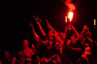 A group of people is gathered at a lively event, with several individuals holding up lit flares and others capturing the scene on their phones. The atmosphere is vibrant and celebratory, with a red hue illuminating the participants.
