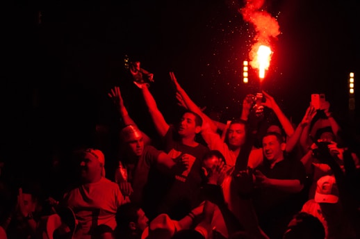 A group of people is gathered at a lively event, with several individuals holding up lit flares and others capturing the scene on their phones. The atmosphere is vibrant and celebratory, with a red hue illuminating the participants.