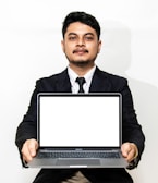 A person is holding a laptop with a blank white screen, dressed in formal attire including a suit, tie, and white shirt. The background is plain and white.