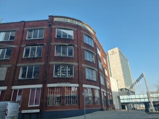 A multi-story brick building with numerous windows, featuring the name 'JACOBS AND CO CARDIFF' at the top. Another building, the 'Clayton Hotel,' stands in the background with a modern design. The street in front has a few parked vehicles, and a railway bridge is visible to the right.