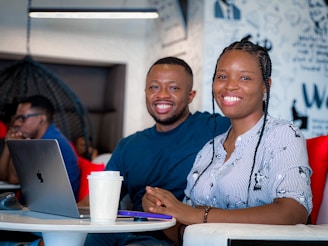 A man and a woman sit side by side, both smiling at the camera. They are in a modern workspace with a laptop and a coffee cup on the table in front of them. The background has wall art and other people working.