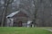 A rustic, weathered log cabin with red-framed windows stands amid a sparse forest of leafless trees. The cabin has a gabled roof made of metal that appears aged and fits the era of the building. A smaller structure is attached to the cabin's side. The surrounding ground is covered with patchy green grass, while bare trees dominate the background, creating a sense of stillness.